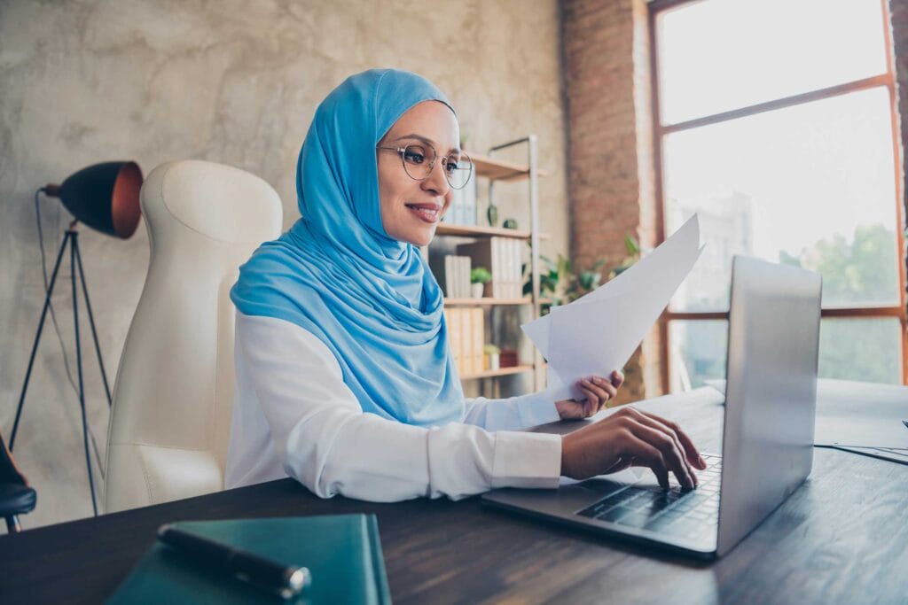 A muslim woman holding a document to translate while using a laptop