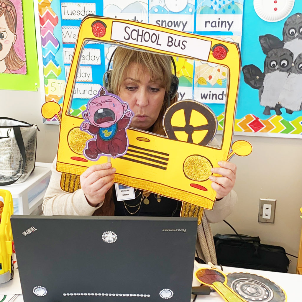 A teacher uses a paper school bus and puppets to relate the song "Wheels on the Bus" to newcomer children.