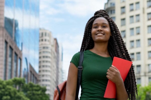 A female refugee youth smiling in the city carrying a bag and a folder