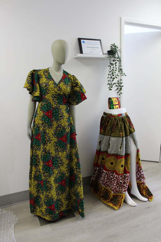 A yellow, red and green dress on a mannequin. To the right, a yellow, red, white and black dress on the bottom half of a mannequin. A red, yellow, and green purse sits on top of the half of a mannequin 