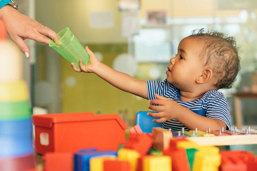 A child surrounded by toys reaching out for a cup given by a woman