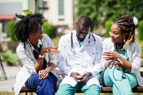 Three African American group doctors with stethoscope wearing lab coat sitting on bench.