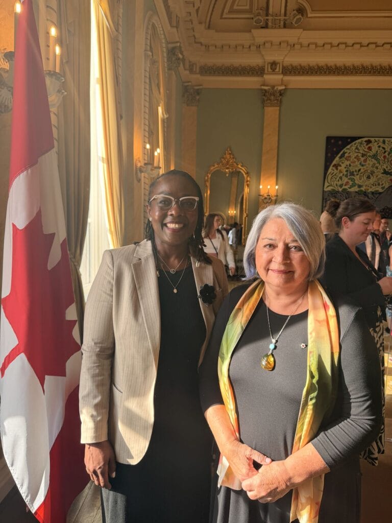 Two women posing for a photo in an ornate room with a Canadian flag nearby, smiling at the camera.