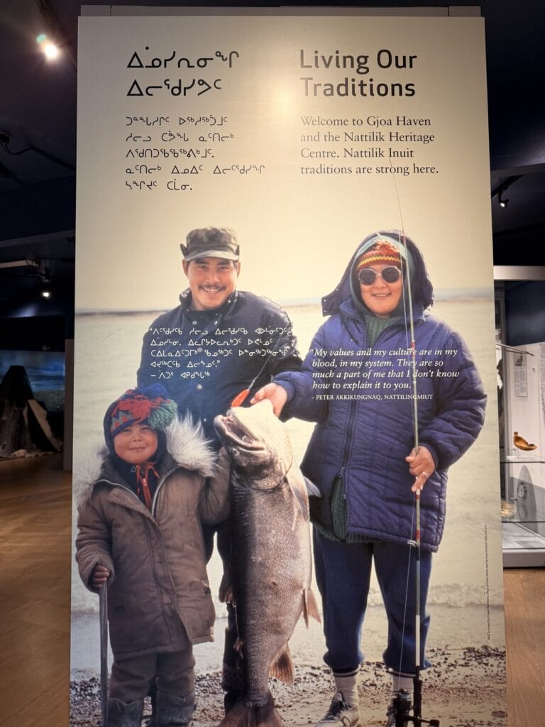 Family of three Inuit people posing on a beach with a large fish, under a banner that says "Living Our Traditions."