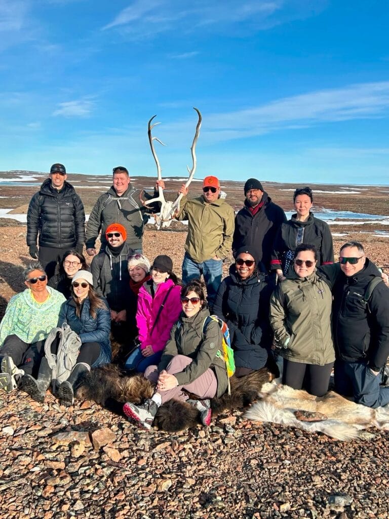 Group of hikers posing outdoors on rocky tundra, several people holding large antlers in a clear blue sky.