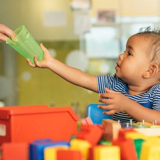 A child surrounded by toys reaching out for a cup given by a woman