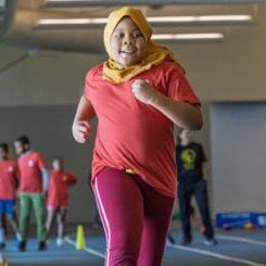 Girl wearing a yellow hijab and red shirt runs on an indoor track, with teammates in red shirts in the background.