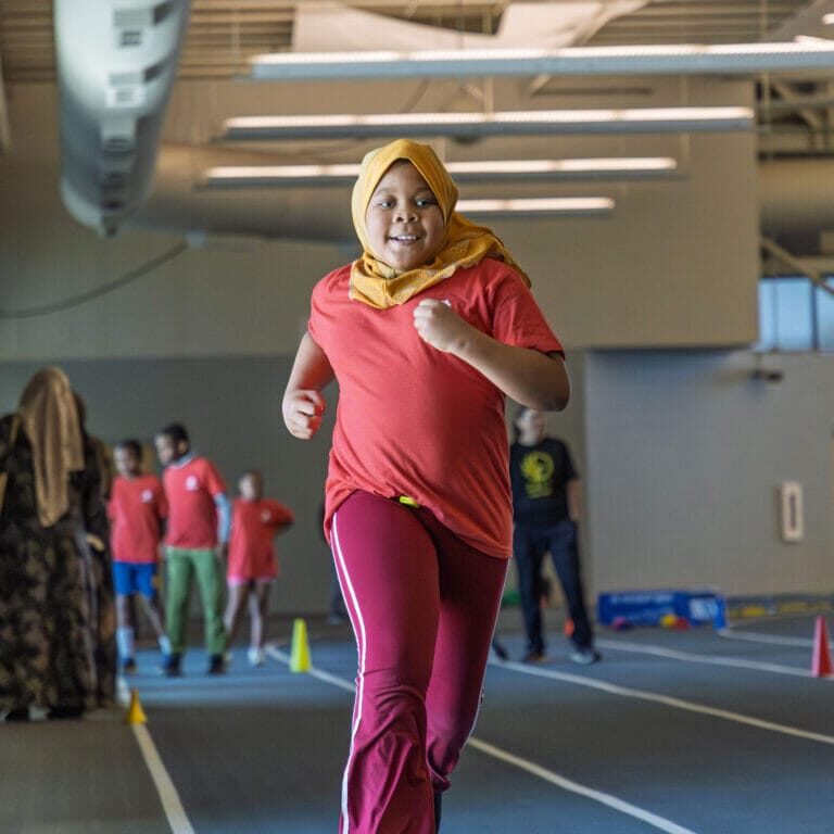 Girl wearing a yellow hijab and red shirt runs on an indoor track, with teammates in red shirts in the background.