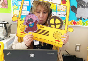 A teacher uses a paper school bus and puppets to relate the song "Wheels on the Bus" to newcomer children.