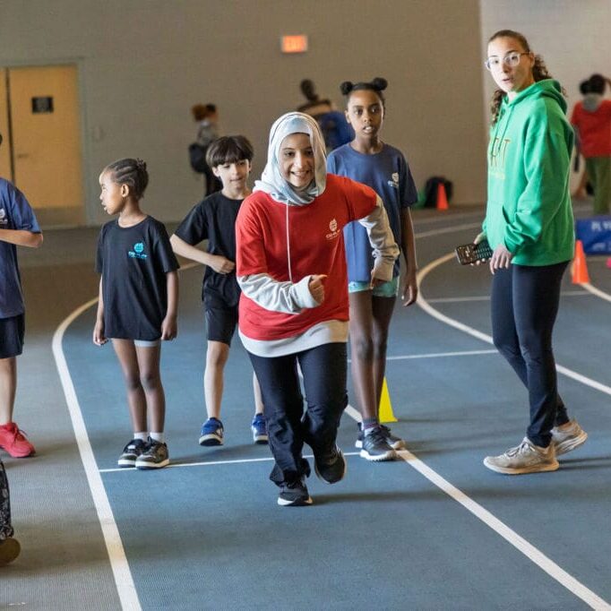 Children in colorful shirts run on an indoor track, while a woman in a green hoodie watches nearby and spectators clap on the left edge.
