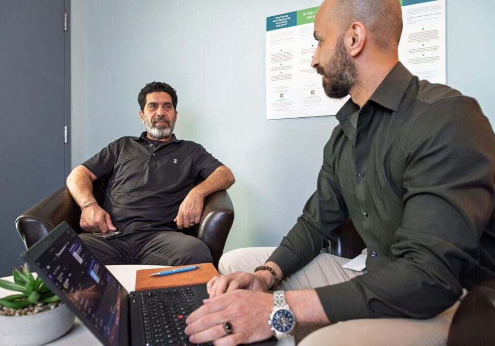 Two men in a meeting: one relaxed in a chair with a laptop, the other leaning in and listening in an office setting.