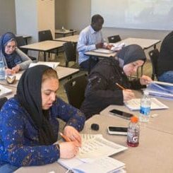 Several adult students sit at desks in a classroom, writing in notebooks; a Canadian flag on the wall and a whiteboard in the background suggest a learning session.