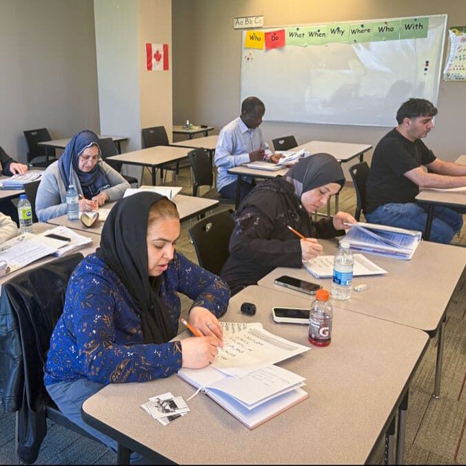 Several adult students sit at desks in a classroom, writing in notebooks; a Canadian flag on the wall and a whiteboard in the background suggest a learning session.