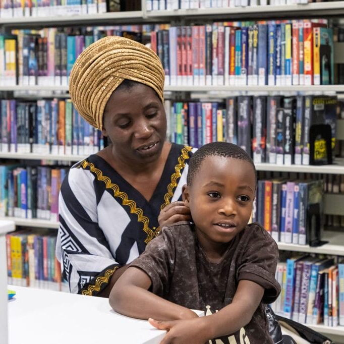 Woman wearing a gold head wrap sits with a young boy at a white table in a library. The colorful bookshelves loom in the background on shelves behind them.