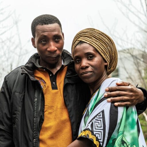 Couple posing outdoors with arms around each other; man in black jacket and orange polo, woman in a gold head wrap and green scarf.