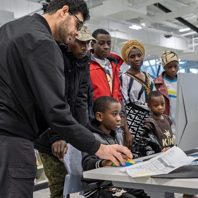 Man and four children gathered around a computer, examining printed worksheets in a bright library or classroom setting.