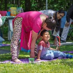 A woman in a pink outfit leaning toward a smiling boy on a purple mat at an outdoor gathering in a park.