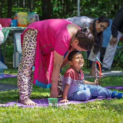 A woman in a pink outfit leaning toward a smiling boy on a purple mat at an outdoor gathering in a park.