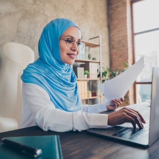 A muslim woman holding a document to translate while using a laptop