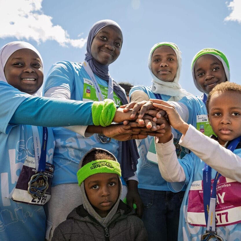 Group of smiling volunteers in blue shirts join hands in a circle outdoors, wearing race bibs and medals against a bright blue sky.
