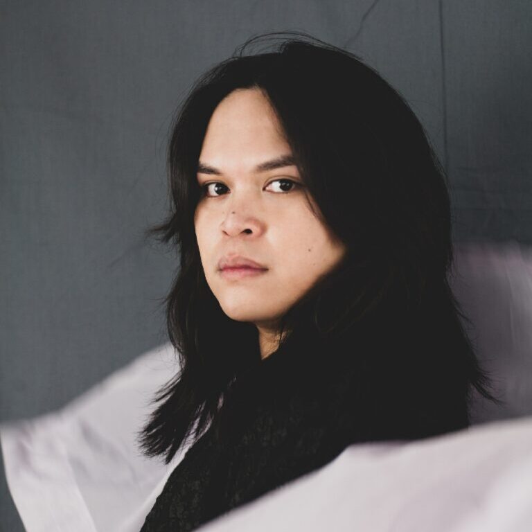 A portrait of Excel Garay, an artist. She has black hair wearing black clothe looking towards the camera while standing sideways.